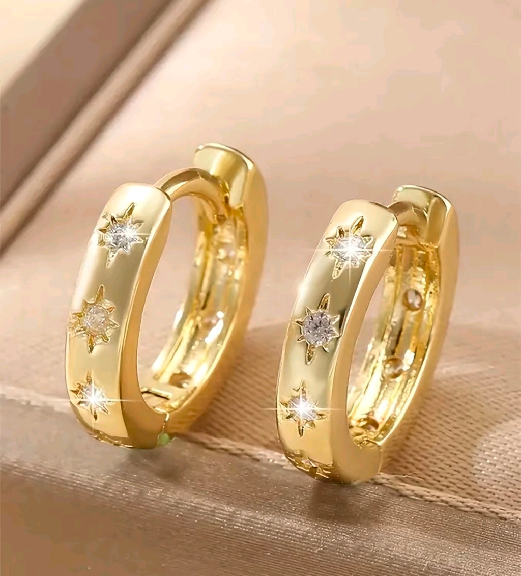 Argollas doradas pequeñas y gruesas que quedan bien pegadas a la oreja. Tienen tres estrellas grabadas al frente y cada una lleva una piedra brillante en el centro. Se abren y cierran con una bisagra trasera.