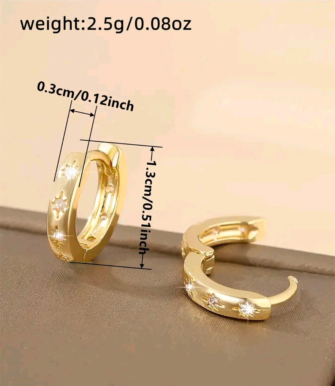 Argollas doradas pequeñas y gruesas que quedan bien pegadas a la oreja. Tienen tres estrellas grabadas al frente y cada una lleva una piedra brillante en el centro. Se abren y cierran con una bisagra trasera.