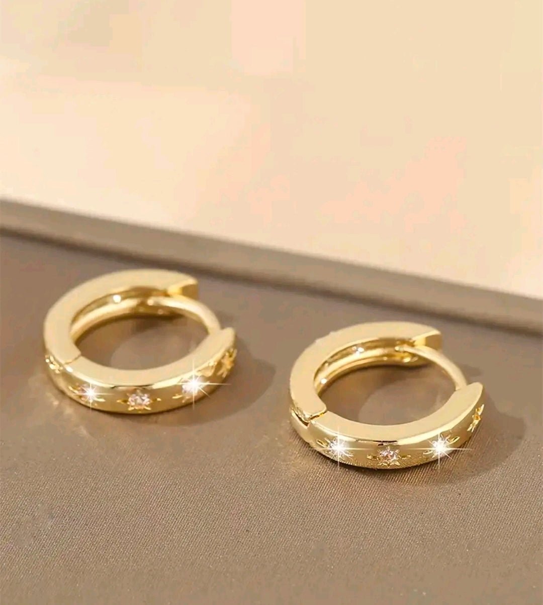 Argollas doradas pequeñas y gruesas que quedan bien pegadas a la oreja. Tienen tres estrellas grabadas al frente y cada una lleva una piedra brillante en el centro. Se abren y cierran con una bisagra trasera.