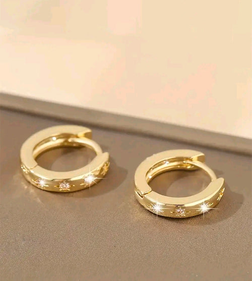 Argollas doradas pequeñas y gruesas que quedan bien pegadas a la oreja. Tienen tres estrellas grabadas al frente y cada una lleva una piedra brillante en el centro. Se abren y cierran con una bisagra trasera.